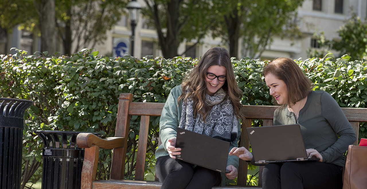 Two graduate students working on laptops sitting on a bench on campus.