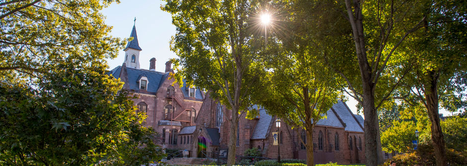 An image of President's Hall and the sun shining through trees above the chapel. 
