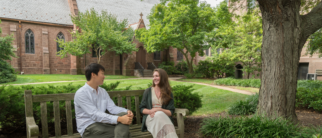 Image of two students sitting on a bench with a chapel in the background.